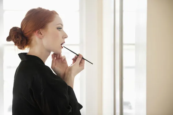 Vista de perfil de una mujer joven en bata aplicando delineador de labios —  Fotos de Stock