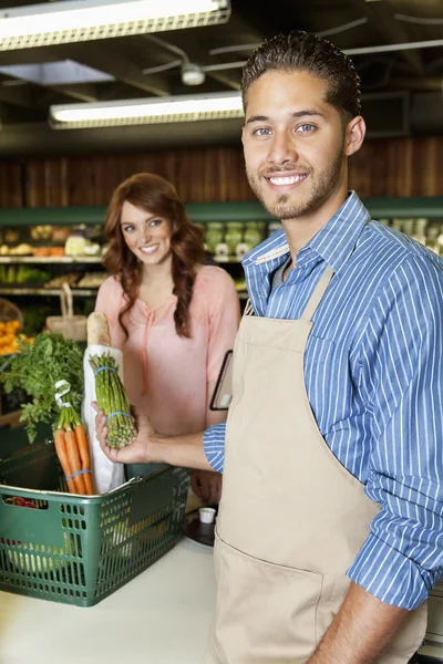 Ritratto di un giovane commesso felice che tiene verdure con donna sullo sfondo — Foto Stock