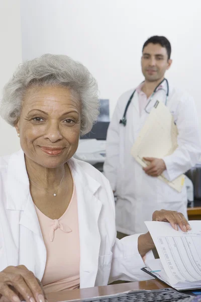 Doctor sentado con documentos y colegas de pie en segundo plano —  Fotos de Stock