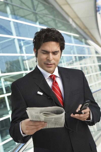 Бизнесмен читает газеты, пользуясь мобильным телефоном — стоковое фото