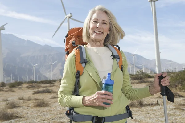 Senior vrouw met wandelen paal en rugzak op windmolenpark — Stockfoto