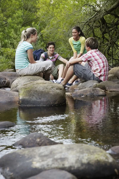 Teenage boys and girls sitting on stones by river in forest — Stock Photo, Image