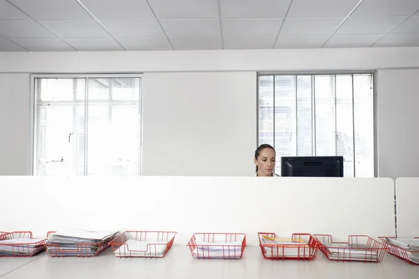 Female office worker using computer in office cubicle behind trays with documents