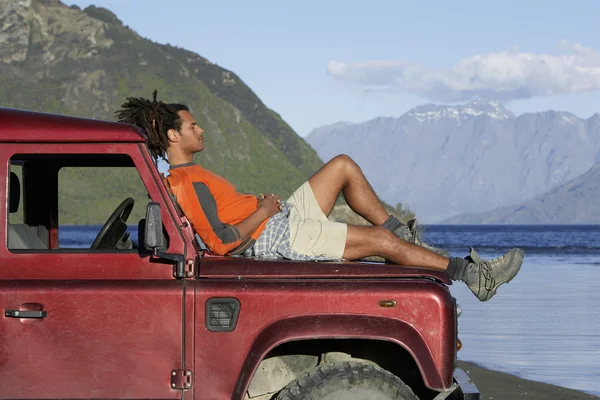 Uomo sdraiato sul cappuccio della jeep vicino al lago di montagna — Foto Stock