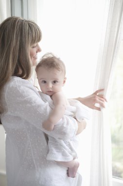 Anne erkek bir bebek tutarken pencereden bakarak duruyor