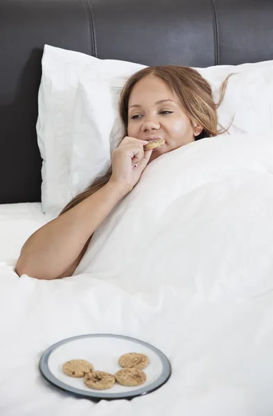 Pregnant woman eating cookies in bed — Stock Photo, Image