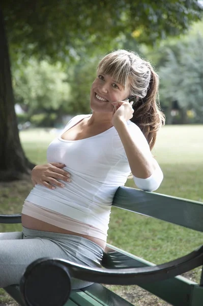 Беременная женщина на скамейке в парке на мобильном телефоне — стоковое фото