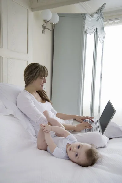 Mãe senta-se digitando no laptop com bebê ao lado dela na cama — Fotografia de Stock