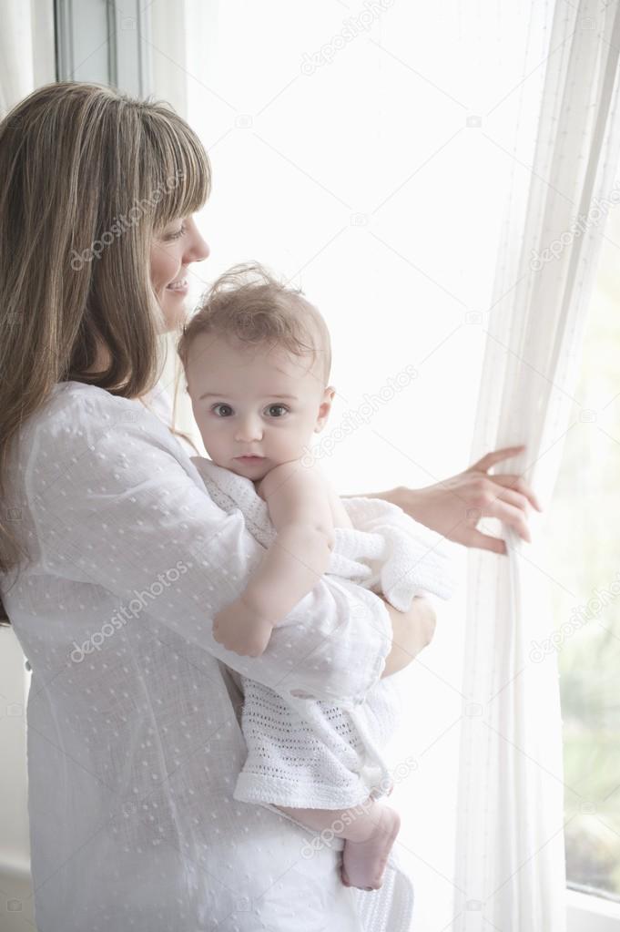 Mother stands looking through window while holding a baby boy