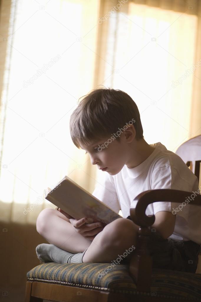 Boy sits cross-legged on chair reading book
