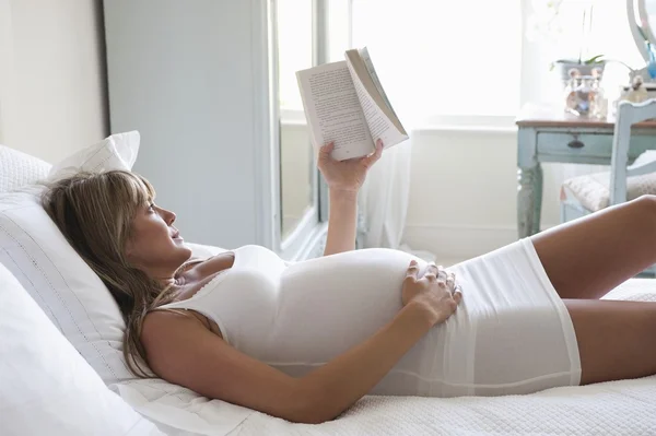 Беременная женщина лежит на кровати, читая книгу — стоковое фото