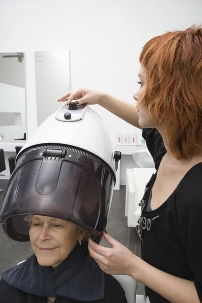 Eldelrly žena sedí pod kapotou hairdrying — Stock fotografie