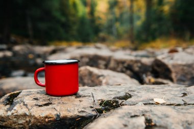 Kahveli kırmızı bir kupa Sonbahar sarı ormanında bir nehrin yanındaki bir taşın üzerinde duruyor.