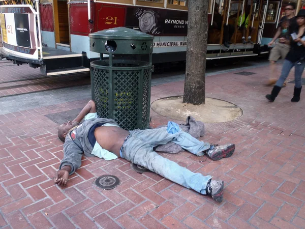 Homeless man sleeps on ground resting against a trash can next t — Stock Photo, Image