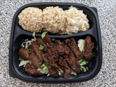 Biftek Katsu, kahverengi pirinç ve tepside siyah plastik tabakta Mac salatası..