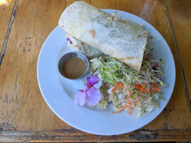 Close-up of Tofu Burrito that been cut in half with salad, dressing, and flower on a plate sitting on old table.