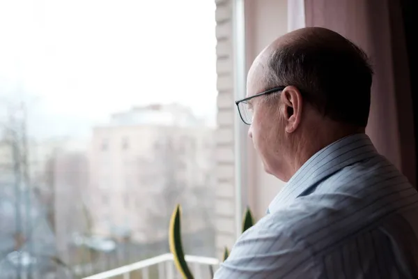 Homme caucasien âgé regardant par la fenêtre avec expression solitaire — Photo