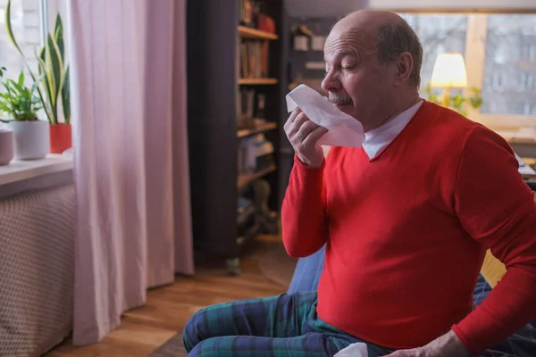 Hombre mayor sosteniendo un pañuelo y estornudando sentado en casa — Foto de Stock