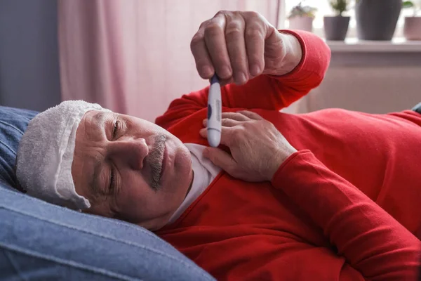 A sick male checking thermometer lying with wet towel on his forehead — Stockfoto
