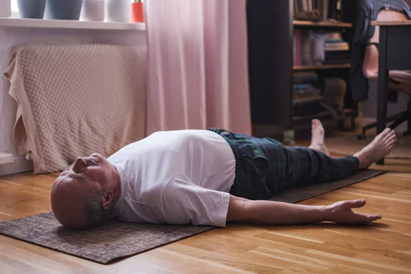 Kıdemli adam ahşap bir zeminde meditasyon yapıyor ve Shavasana pozunda yatıyor..