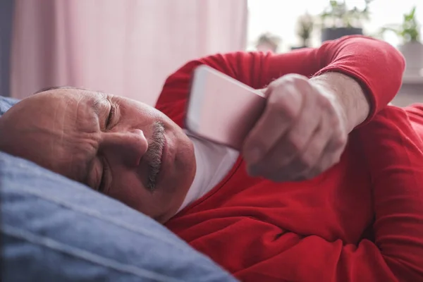 Старший кавказский мужчина с помощью смартфона лежит на диване дома. — стоковое фото