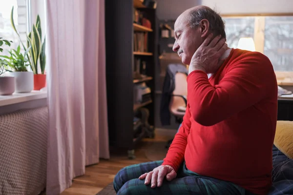Frustrated senior man in red sweater holding hand on his neck sitting on sofa — Stock Photo, Image