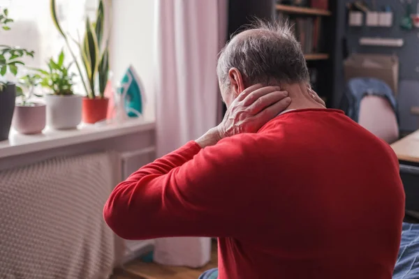Frustrated senior man in red sweater holding hand on his neck sitting on sofa — Stockfoto