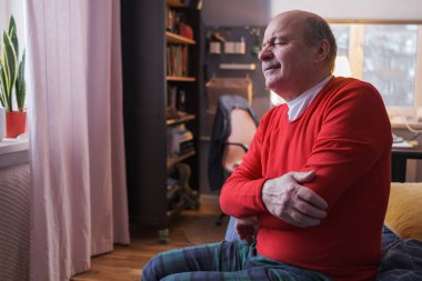 Dirseği ağrıyan yaşlı bir adam evinde kanepede oturuyor..