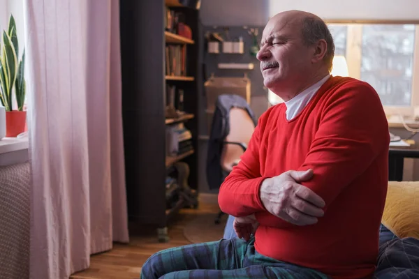 Dirseği ağrıyan yaşlı bir adam evinde kanepede oturuyor..