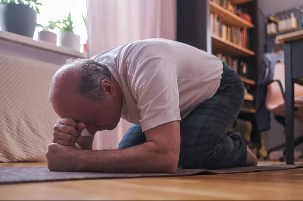 Senior man doing the child pose while practicing yoga alone in his living room Imagens De Bancos De Imagens