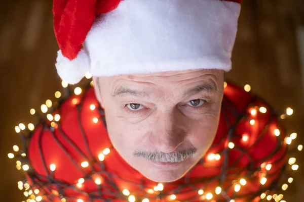 Senior homme triste en chapeau de Noël rouge enveloppé dans la guirlande. — Photo