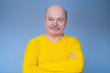 Kıdemli adam kameraya gülümserken çapraz ellerle poz veriyor.