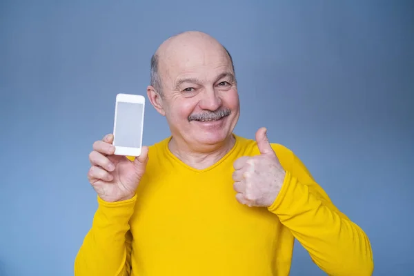 身穿黄色毛衣的英俊的老年男子露出大拇指，并在电话屏幕上显示出来 — 图库照片
