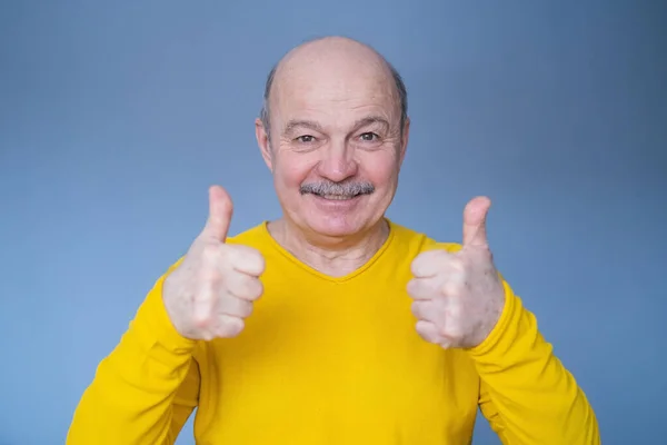 Przystojny starszy pan w żółtym swetrze pokazuje kciuk do góry. — Zdjęcie stockowe