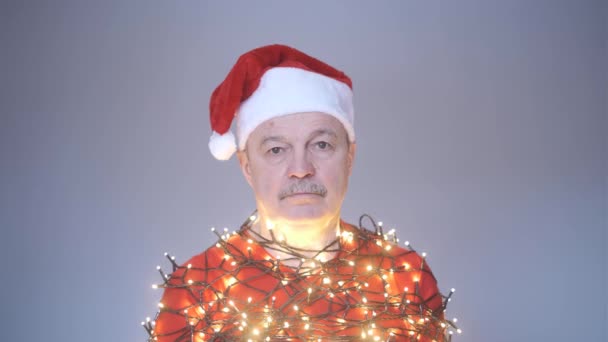 Старший сумний чоловік у червоному різдвяному капелюсі, загорнутий у гірлянду . — стокове відео
