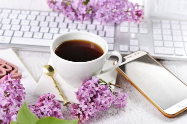 Bir fincan kahve, akıllı telefon ve üzerinde mor leylak çiçekleri olan beyaz klavye. 