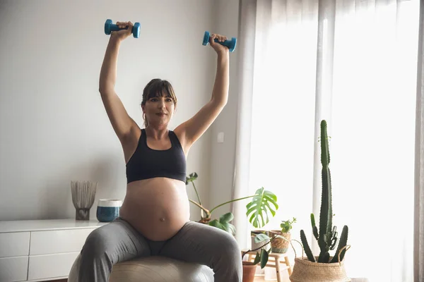 ダンベルと仕事をしている妊婦は 自宅でフィットボールで筋力トレーニングをしています 赤ちゃんを待っている間 良い形を保ちます — ストック写真