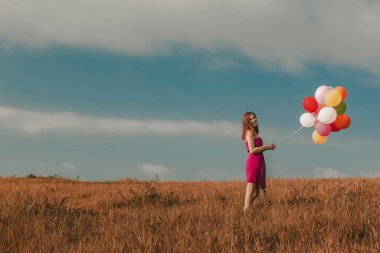 Güzel genç bir kadın elinde balonlarla çayırda yürüyor.