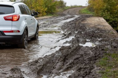Kirli yolda ilerleyen temiz beyaz SUV aracı. Bulanık yamacın önünde ıslak kil var.