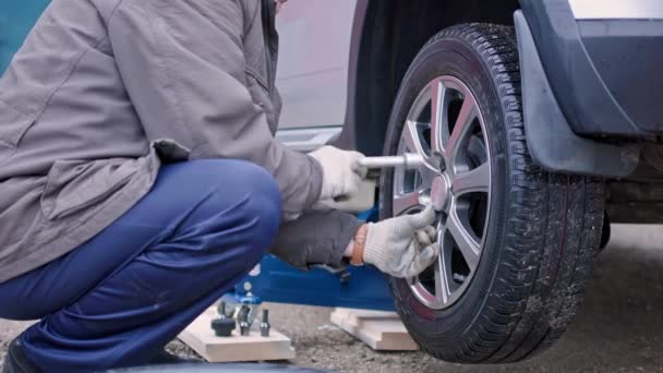 Starý muž ve špinavých kalhotách odstranění pneumatiky stříbrného auta v zamračený podzimní den — Stock video