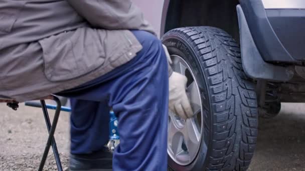 Invecchiato uomo in pantaloni blu imposta inverno tempestato di pneumatico d'argento auto a nuvoloso giorno d'autunno — Video Stock