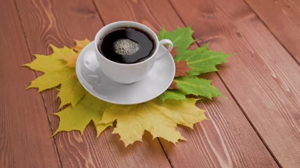 Φλιτζάνι καφέ σε ξύλινο τραπέζι με πολύχρωμα φθινοπωρινά φύλλα σφενδάμου με περιστρεφόμενες φυσαλίδες καφέ — Αρχείο Βίντεο