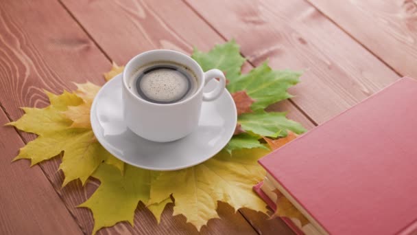 Xícara de café na mesa de madeira com livro e folhas de bordo outonal colorido com bolhas de café girando — Vídeo de Stock
