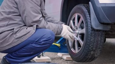 Kirli mavi pantolonlu ve gri ceketli yaşlı adam gümüş arabadaki vidaları söküyor.