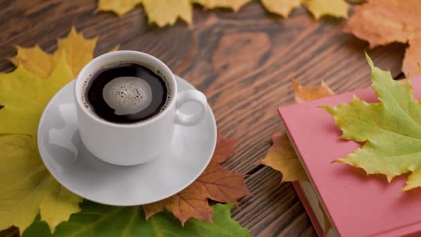 Coffe kopp på träbord med bok och färgglada höstliga lönnlöv med snurrande kaffebubblor — Stockvideo