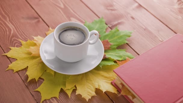 Φλιτζάνι καφέ σε ξύλινο τραπέζι με βιβλίο και πολύχρωμα φθινοπωρινά φύλλα σφενδάμου με περιστρεφόμενες φυσαλίδες καφέ — Αρχείο Βίντεο