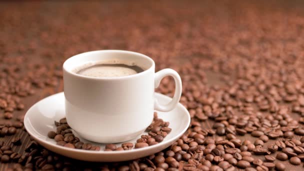 Café negro en taza blanca pequeña en el plato con burbujas giratorias en una superficie plana cubierta con granos de café tostados — Vídeo de stock