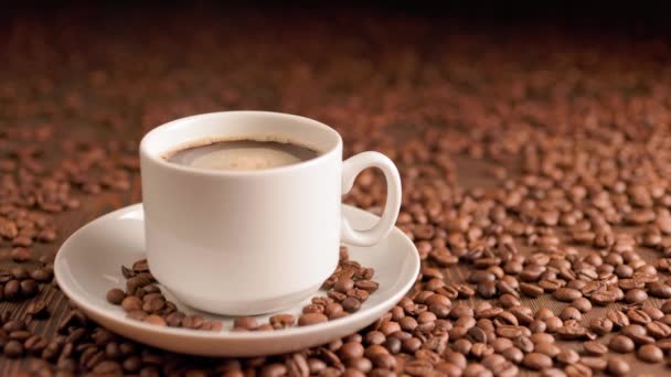 Черный кофе в маленькой белой кружке на тарелке с вращающимися пузырьками на плоской поверхности, покрытой жареной кофейной зёрной — стоковое видео