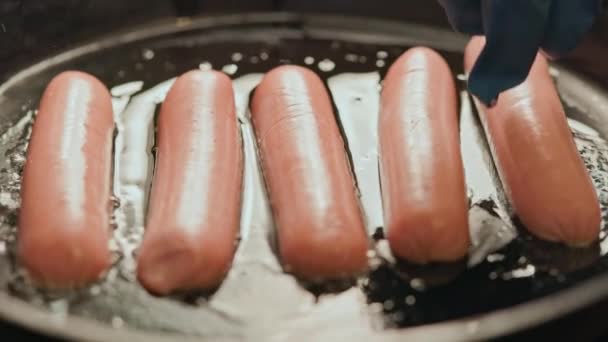 Рука в голубой перчатке, перекладывающая сосиски в горячую сковородку, покрытую подсолнечным маслом — стоковое видео