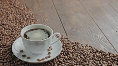 Düz ahşap bir yüzey üzerinde dönen baloncuklar bulunan beyaz bir fincanda koyu kahve kısmen kavrulmuş kahve çekirdekleri ile kaplı.